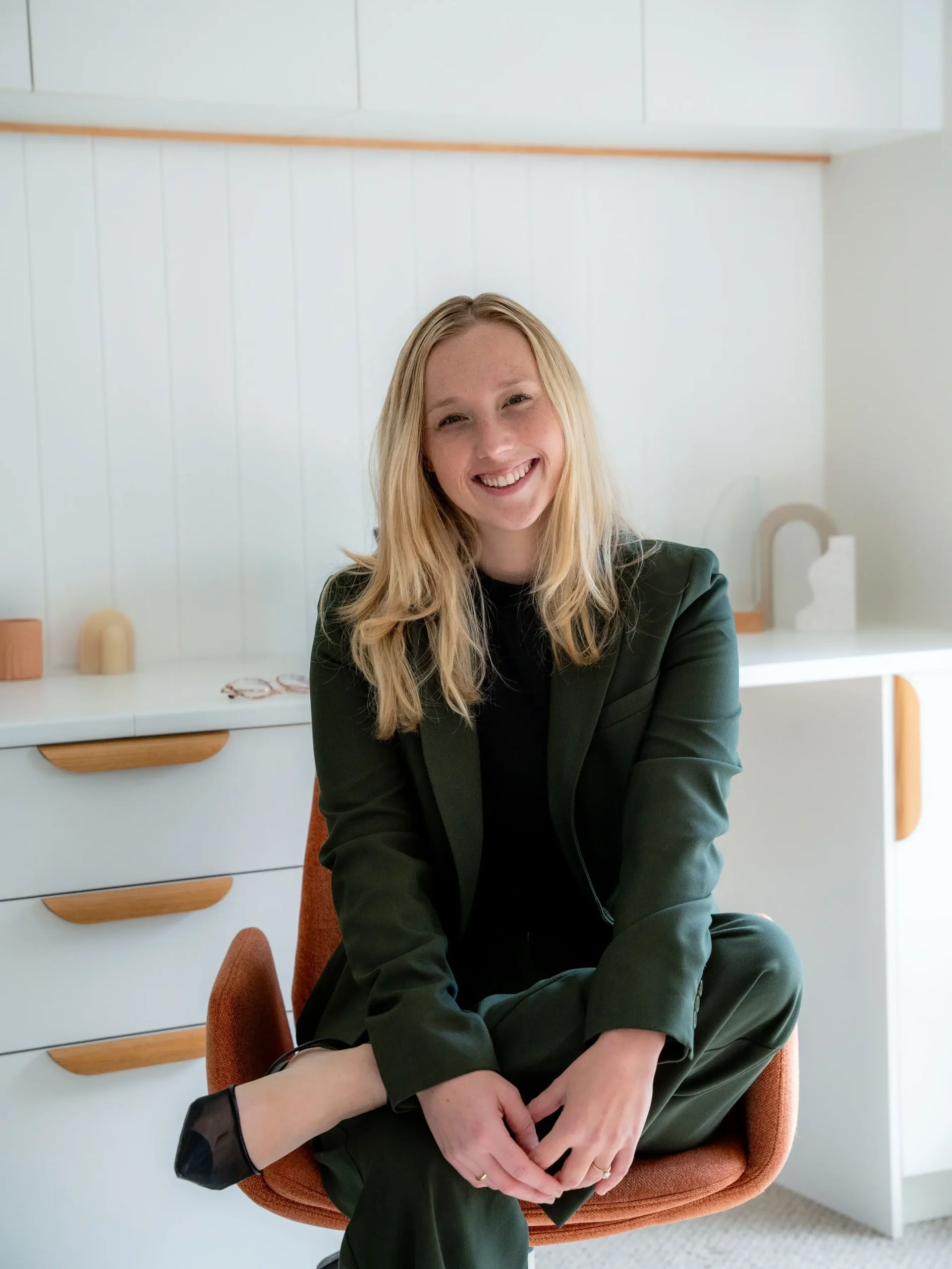 Smiling blonde woman wearing a dark green pantsuit sitting cross-legged in an orange desk chair inside a bright office setting.