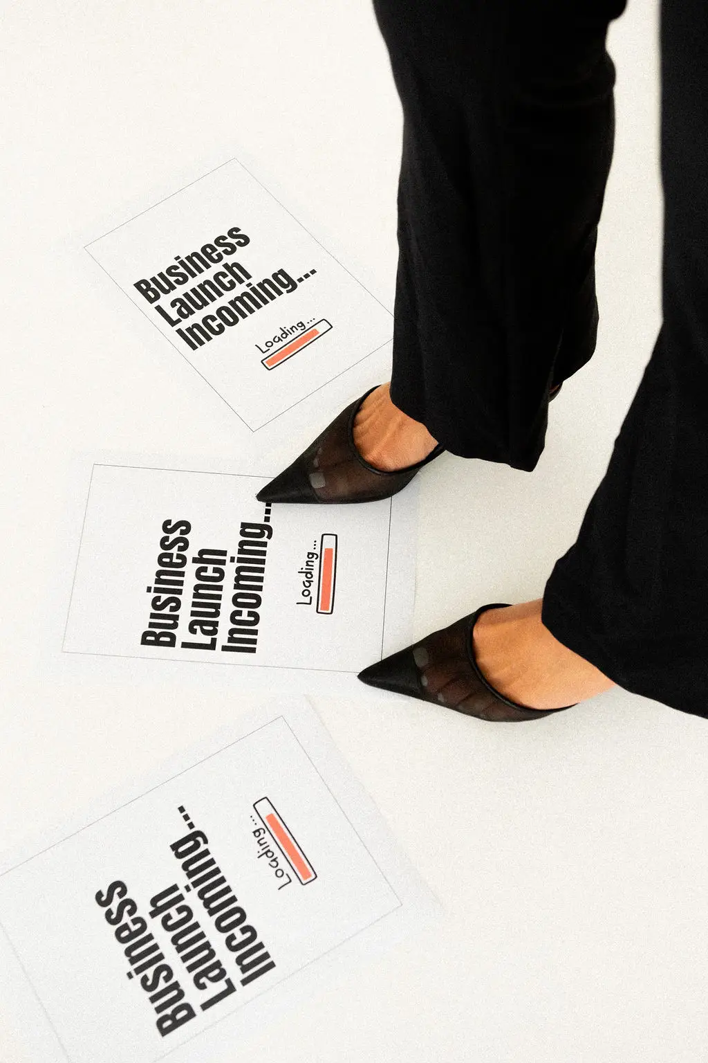 High-angle shot of a person in black trousers and pointed heels standing over printed signs reading Business Launch Incoming Loading on a white floor.