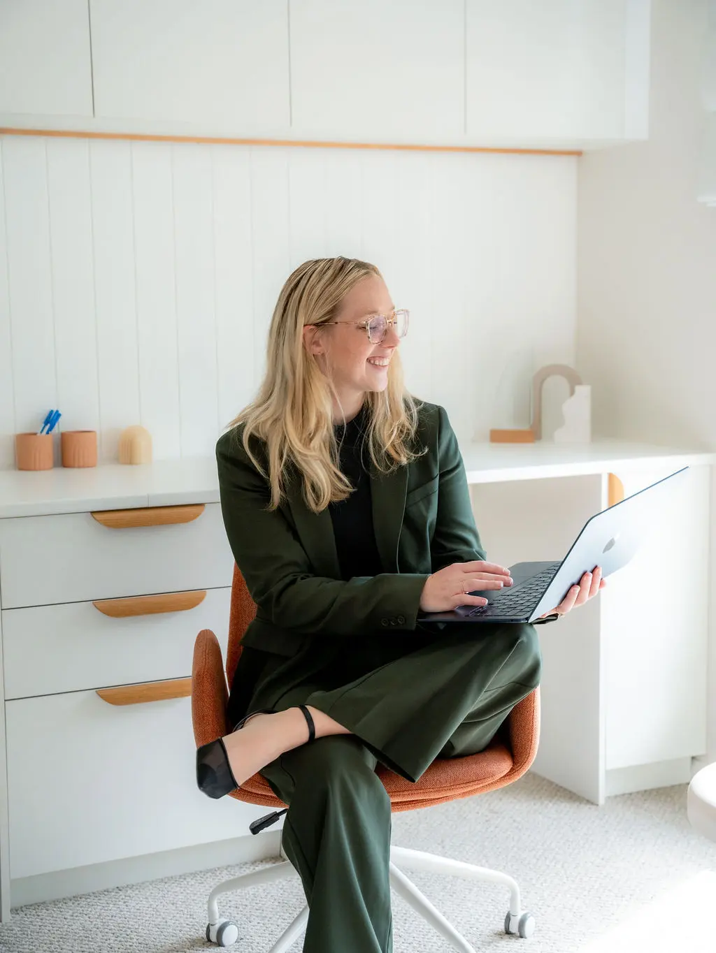 A smiling blonde woman in a dark green suit and glasses sitting cross-legged in an orange desk chair, holding a laptop in a bright, white office.