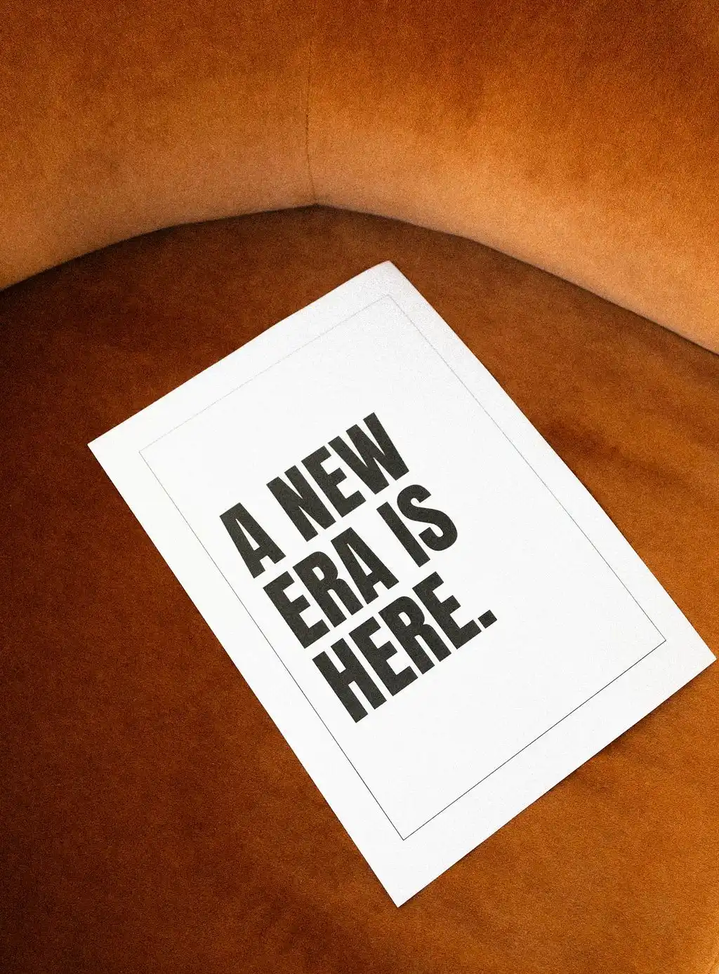 Top-down view of a white printed card reading A New Era Is Here resting on a textured orange velvet armchair.